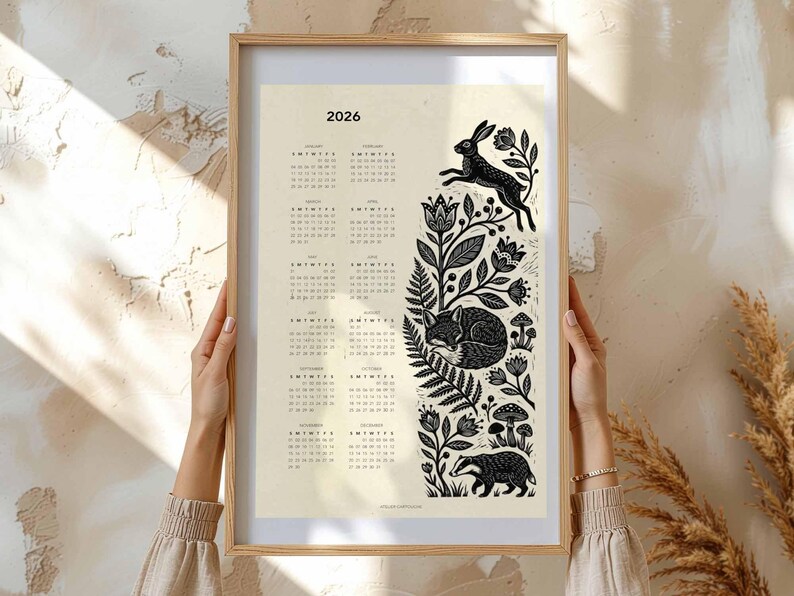 May include: A framed 2026 calendar print with a cream background. The calendar features a black and white illustration of a rabbit, fox, and badger among floral and botanical elements. The frame is a light wood color.