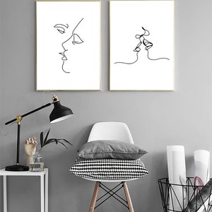 May include: Two framed minimalist line art prints. One depicts a side profile of a face, the other shows two faces kissing. The art is black on a white background, framed in gold, and displayed against a gray wall.