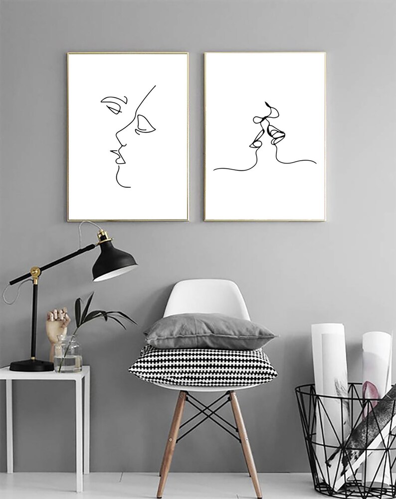 May include: Two framed minimalist line art prints. One depicts a side profile of a face, the other shows two faces kissing. The art is black on a white background, framed in gold, and displayed against a gray wall.
