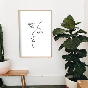 May include: A framed minimalist art print featuring a continuous line drawing of two faces in profile, appearing to kiss. The artwork is in black against a white background, set within a light wood frame. The print is displayed on a white wall.