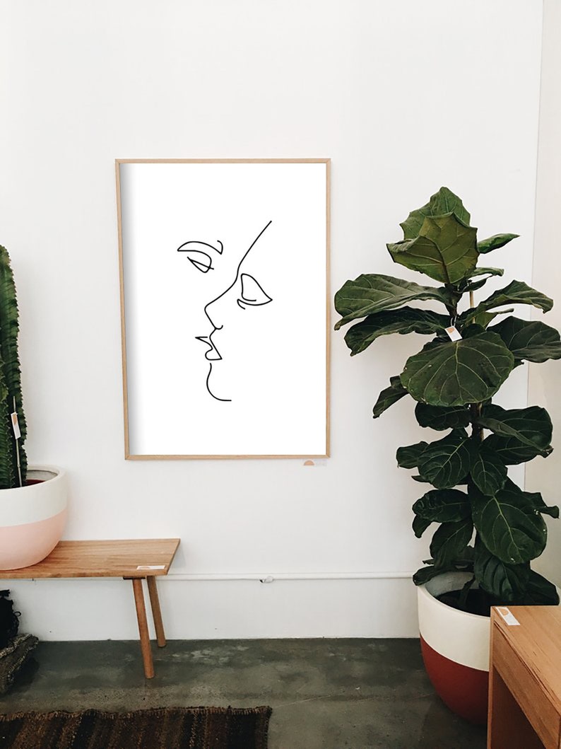 May include: A framed minimalist art print featuring a continuous line drawing of two faces in profile, appearing to kiss. The artwork is in black against a white background, set within a light wood frame. The print is displayed on a white wall.