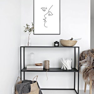 May include: A black framed print featuring a minimalist line drawing of two faces in profile, kissing. The artwork hangs above a black metal shelf unit with decorative items, including a woven basket and glass jar.