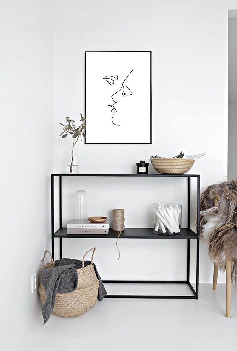 May include: A black framed print featuring a minimalist line drawing of two faces in profile, kissing. The artwork hangs above a black metal shelf unit with decorative items, including a woven basket and glass jar.