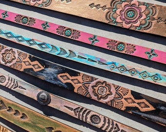 May include: A collection of eight leather belts with various designs, including floral patterns, geometric shapes, and a bull's head. The belts are arranged in a staggered pattern, with the top belt being the most prominent. The belts are all different colors, including brown, pink, blue, and green.
