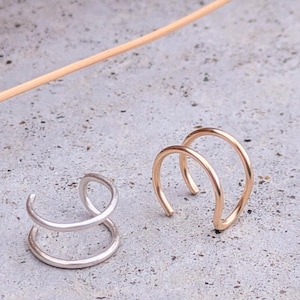 May include: Two ear cuffs, one silver and one gold, are shown on a gray surface. The ear cuffs are made of two thin metal hoops.