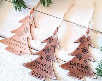 May include: Three Christmas tree ornaments made of light brown leather. Each tree has a unique name and the year 2021 engraved on it. The trees are decorated with snowflakes and stars and are hanging from twine.