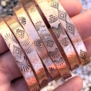 May include: Copper-colored metal bracelets with etched geometric patterns. The bracelets have a hammered texture and are displayed in a hand. The designs include sunbursts, diamonds, and triangles. The bracelets are a warm, metallic color.