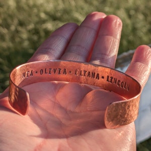 K&ouml;nnte beinhalten: Ein Kupferarmband mit geh&auml;mmerter Oberfl&auml;che und den eingravierten Namen "MIA, OLIVIA, ELIANA, LINCOLN".