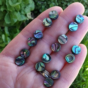 May include: A close-up of a hand holding a collection of iridescent abalone shell earrings. The earrings are round and have a silver backing.