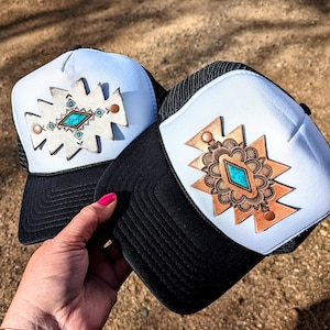 May include: Two white trucker hats with black mesh backs. Each hat features a different leather patch with a turquoise stone and geometric design. One patch is white with brown accents, the other is brown with gold accents.