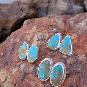 May include: Four pairs of turquoise and silver stud earrings. The earrings feature vibrant turquoise stones with brown and tan matrix patterns, set in polished silver bezels. The earrings are displayed on a textured, reddish-brown stone surface.