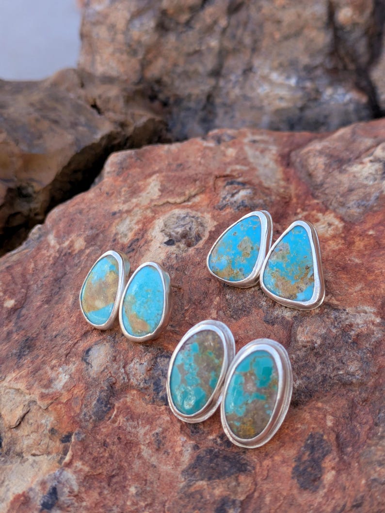 May include: Four pairs of turquoise and silver stud earrings. The earrings feature vibrant turquoise stones with brown and tan matrix patterns, set in polished silver bezels. The earrings are displayed on a textured, reddish-brown stone surface.