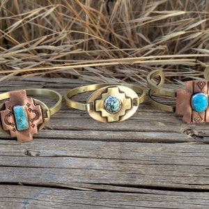 May include: Three brass bracelets with turquoise stones set in copper. Each bracelet features a different design, including a cross, a square, and a diamond pattern.