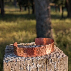 K&ouml;nnte beinhalten: Ein geh&auml;mmertes Kupferarmband mit rustikalem Finish. Das Armband liegt auf einem verwitterten Holzpfosten.