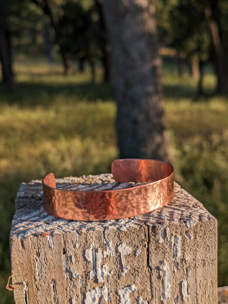 K&ouml;nnte beinhalten: Ein geh&auml;mmertes Kupferarmband mit rustikalem Finish. Das Armband liegt auf einem verwitterten Holzpfosten.