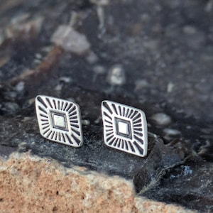 May include: A pair of silver earrings with a geometric design. The earrings are shaped like a diamond with a smaller diamond in the center. The earrings are on a dark background.