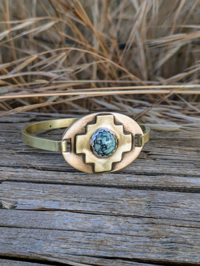 May include: A brass bracelet with a turquoise stone set in a geometric design. The bracelet is on a wooden surface.