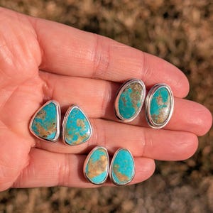 May include: Three pairs of turquoise stud earrings with silver settings. The turquoise stones have a mix of blue and brown hues. The earrings are in oval, teardrop, and triangular shapes.