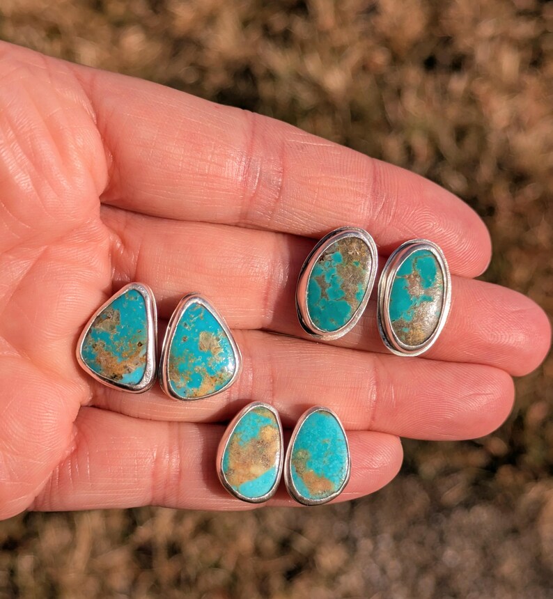 May include: Three pairs of turquoise stud earrings with silver settings. The turquoise stones have a mix of blue and brown hues. The earrings are in oval, teardrop, and triangular shapes.