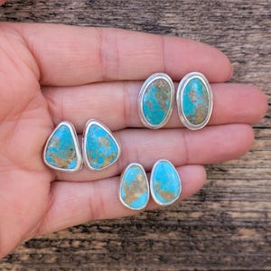 May include: A collection of turquoise stud earrings in various shapes: triangular, oval, and teardrop. Each earring features a vibrant blue-green stone with brown matrix set in a silver bezel. The earrings are displayed on a wooden surface.