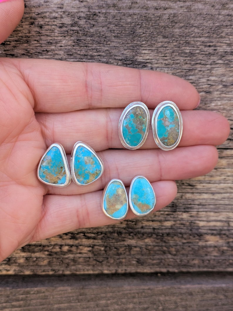 May include: A collection of turquoise stud earrings in various shapes: triangular, oval, and teardrop. Each earring features a vibrant blue-green stone with brown matrix set in a silver bezel. The earrings are displayed on a wooden surface.