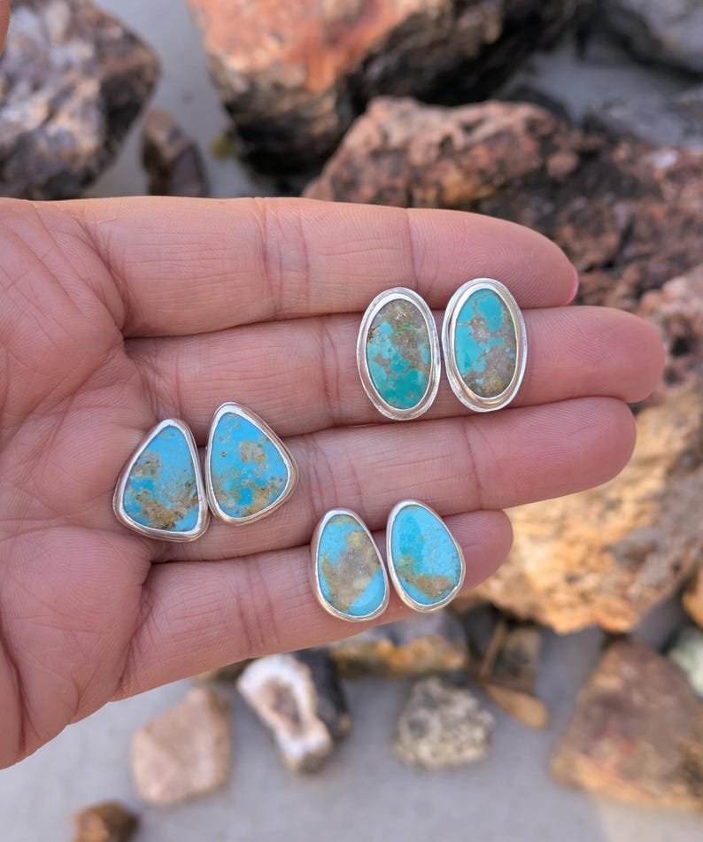 May include: Four pairs of turquoise stud earrings with silver settings. The earrings feature various shapes, including oval and triangular designs. The turquoise stones have a mix of blue and brown hues, and the silver settings have a polished finish.