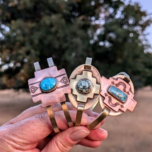 May include: Three brass and copper bracelets with turquoise stones. Each bracelet features a different design with a turquoise stone in the center.