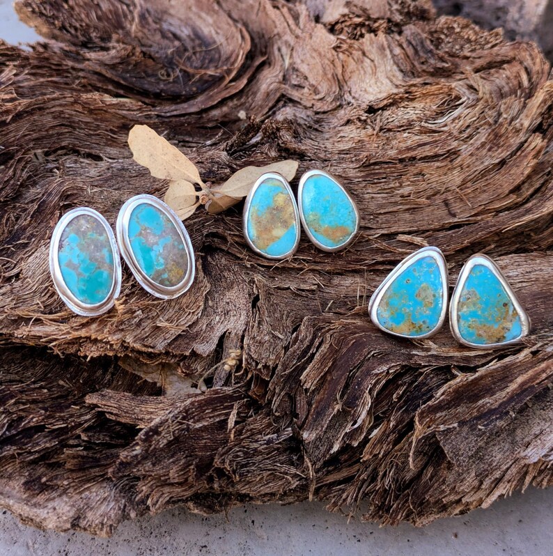 May include: Three pairs of turquoise stud earrings with silver settings. The earrings feature oval, teardrop, and triangular shapes. The turquoise stones have a mix of blue and brown hues, set against a textured wooden background.