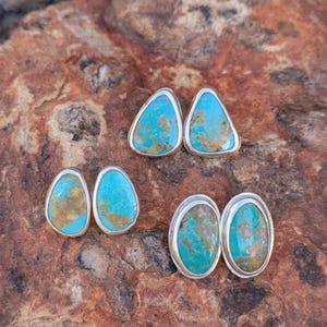 May include: Four pairs of turquoise stud earrings with silver settings. The earrings have a vibrant blue-green color with brown matrix patterns. The earrings are displayed on a textured, reddish-brown stone surface.