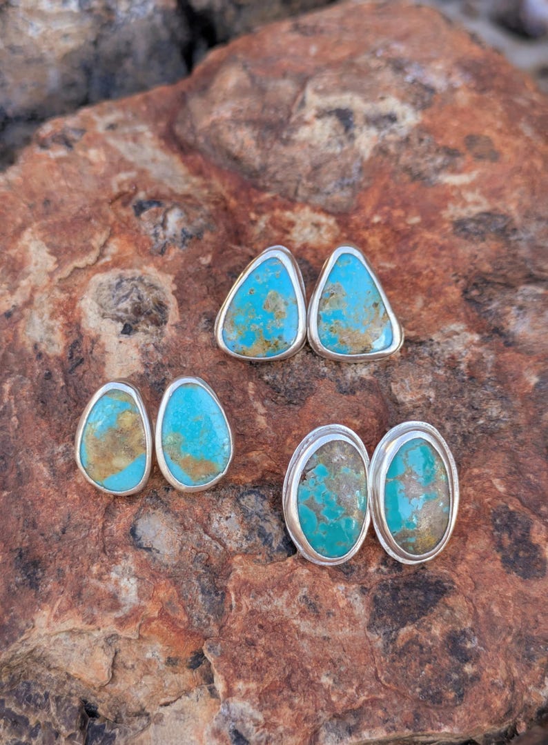 May include: Four pairs of turquoise stud earrings with silver settings. The earrings have a vibrant blue-green color with brown matrix patterns. The earrings are displayed on a textured, reddish-brown stone surface.