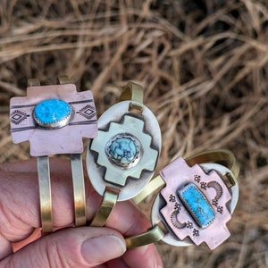 May include: Three brass bracelets with turquoise stones and intricate designs. The bracelets are stacked on top of each other and are held in a hand.