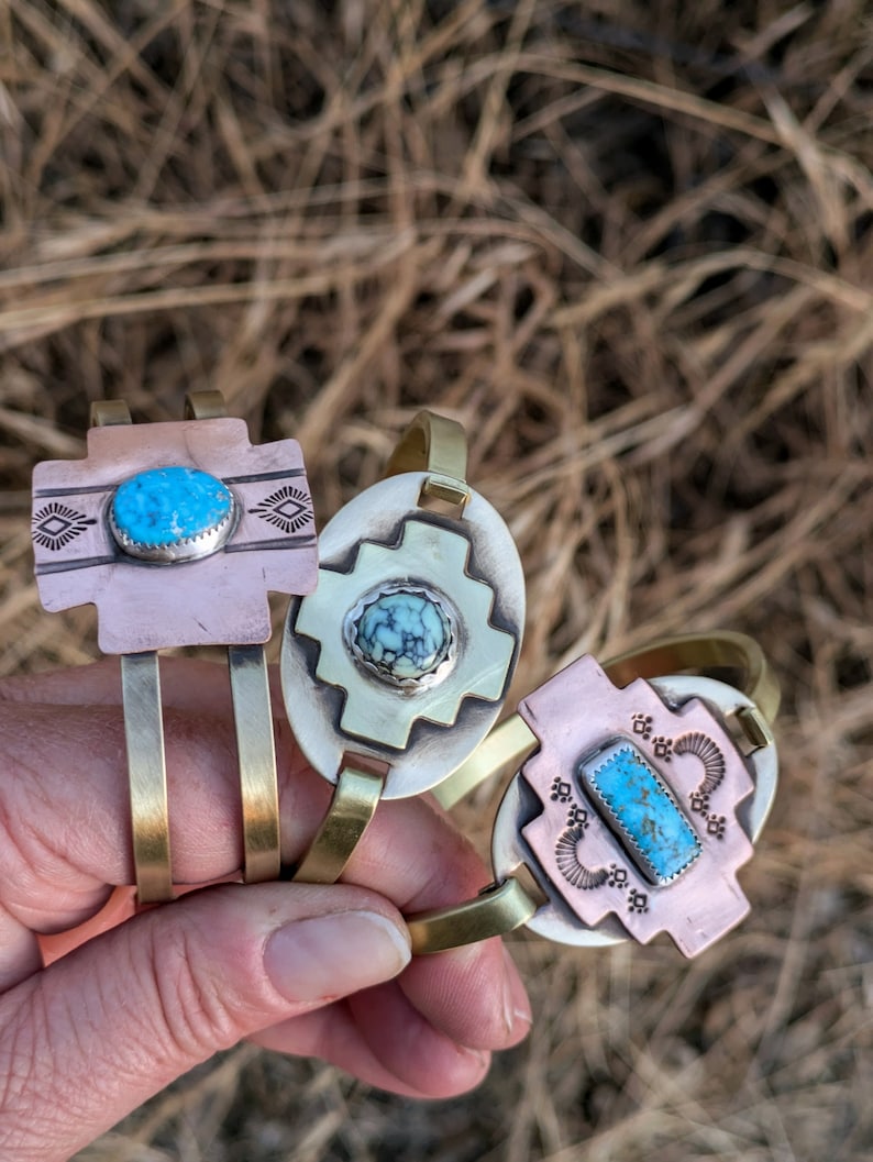 May include: Three brass bracelets with turquoise stones and intricate designs. The bracelets are stacked on top of each other and are held in a hand.