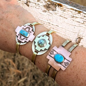May include: Three brass bracelets with turquoise stones set in a cross design. The bracelets are stacked on a person's wrist.