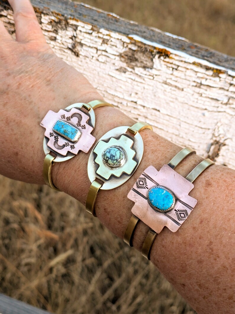 May include: Three brass bracelets with turquoise stones set in a cross design. The bracelets are stacked on a person's wrist.