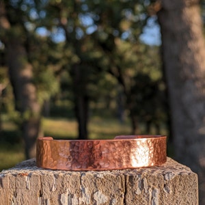 K&ouml;nnte beinhalten: Ein geh&auml;mmertes Kupferarmband mit rustikalem Finish. Das Armband liegt auf einem verwitterten Holzpfosten.