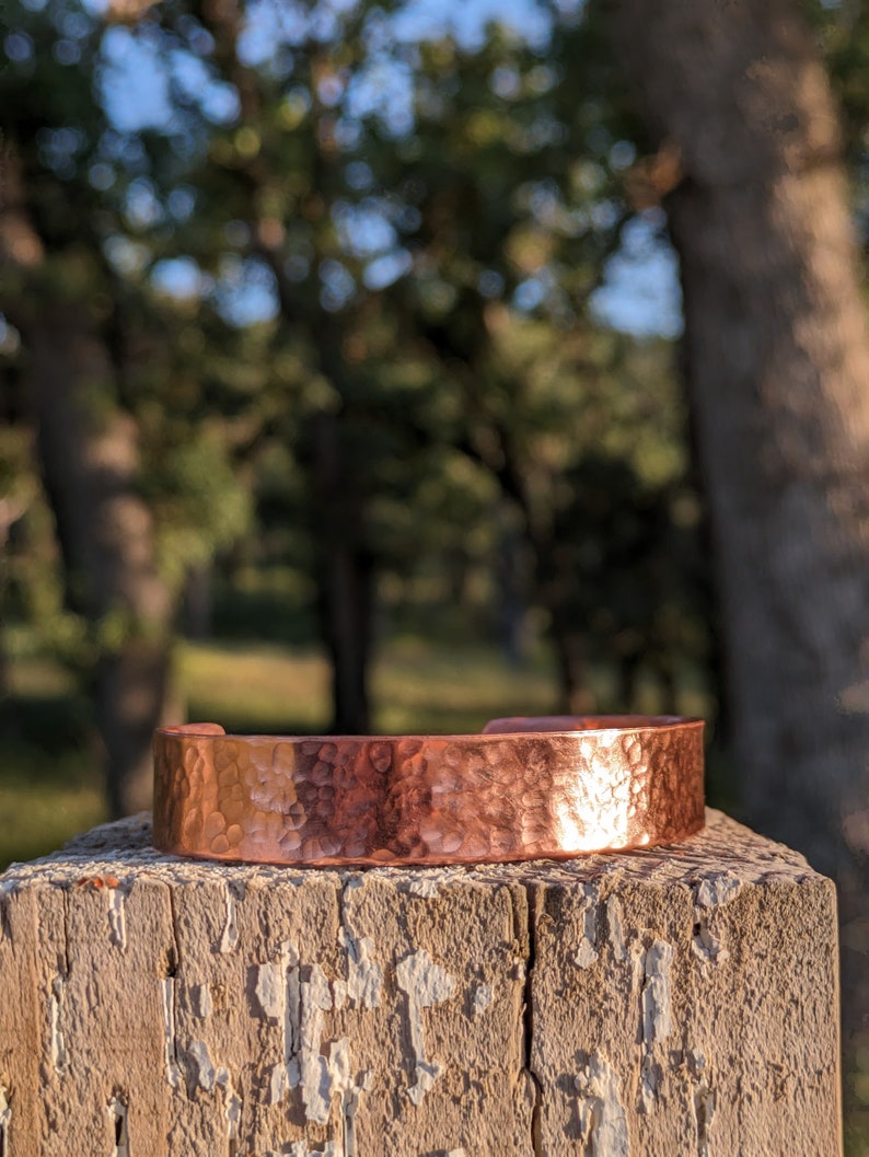 K&ouml;nnte beinhalten: Ein geh&auml;mmertes Kupferarmband mit rustikalem Finish. Das Armband liegt auf einem verwitterten Holzpfosten.