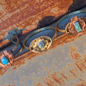 May include: Three brass bracelets with turquoise stones set in copper crosses. The bracelets are arranged on a rusty metal surface.