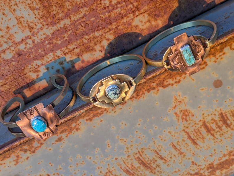 May include: Three brass bracelets with turquoise stones set in copper crosses. The bracelets are arranged on a rusty metal surface.