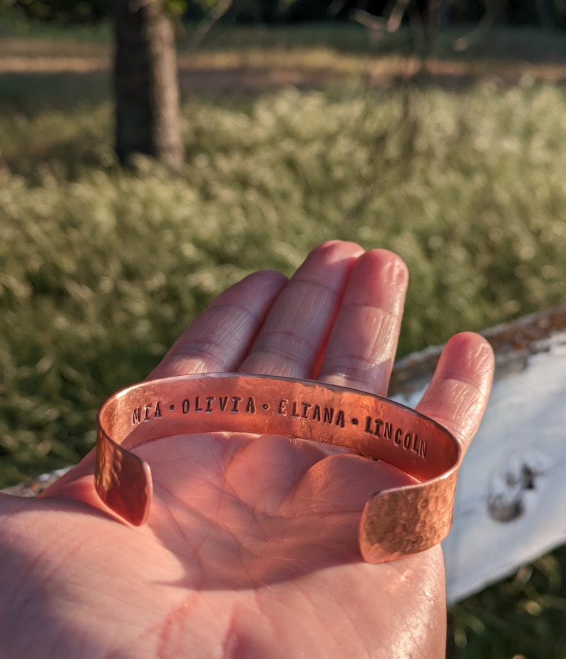 K&ouml;nnte beinhalten: Ein Kupferarmband mit geh&auml;mmerter Oberfl&auml;che und den eingravierten Namen "MIA, OLIVIA, ELIANA, LINCOLN".