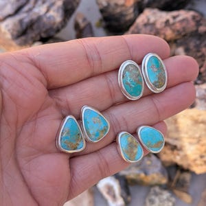 May include: A collection of turquoise stud earrings in various shapes, including oval and teardrop designs. Each earring features a vibrant blue-green stone set in a silver bezel. The natural turquoise stones have brown and tan matrix patterns.
