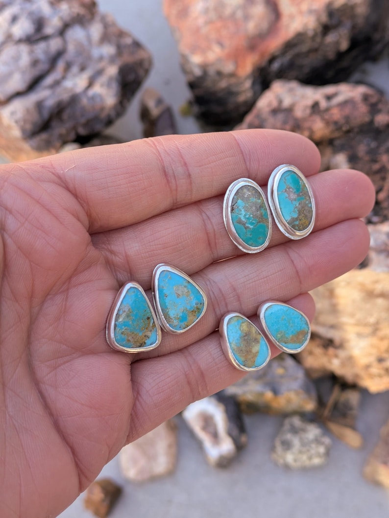 May include: A collection of turquoise stud earrings in various shapes, including oval and teardrop designs. Each earring features a vibrant blue-green stone set in a silver bezel. The natural turquoise stones have brown and tan matrix patterns.