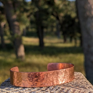 K&ouml;nnte beinhalten: Ein geh&auml;mmertes Kupferarmband mit rustikalem Finish. Das Armband liegt auf einer Holzoberfl&auml;che mit einem verschwommenen Hintergrund aus B&auml;umen.