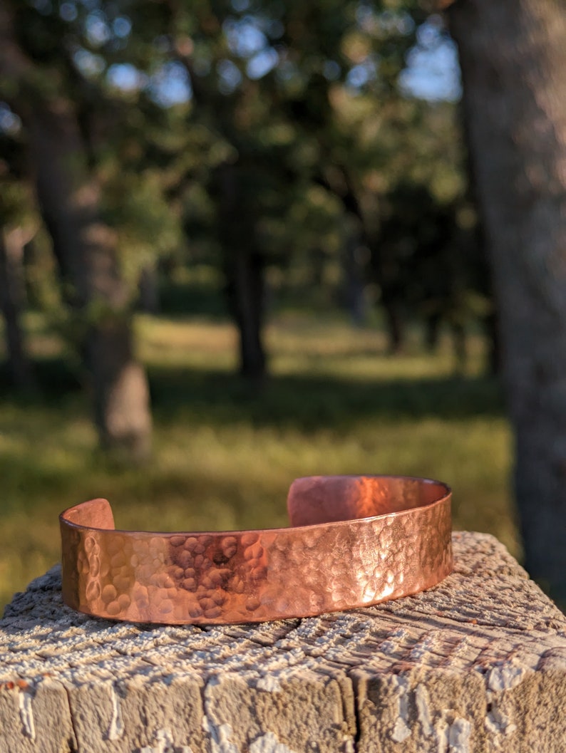 K&ouml;nnte beinhalten: Ein geh&auml;mmertes Kupferarmband mit rustikalem Finish. Das Armband liegt auf einer Holzoberfl&auml;che mit einem verschwommenen Hintergrund aus B&auml;umen.