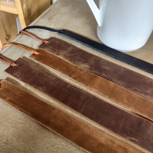 May include: Four leather bookmarks in different shades of brown, with a black leather bookmark in the background. The bookmarks are laid out on a wooden surface.