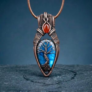 Puede incluir: Un colgante de alambre de cobre enrollado con una piedra de labradorita azul en forma de lágrima y un intrincado diseño de árbol de alambre en su superficie. Una pequeña piedra preciosa ovalada de color rojo anaranjado se sitúa encima.
