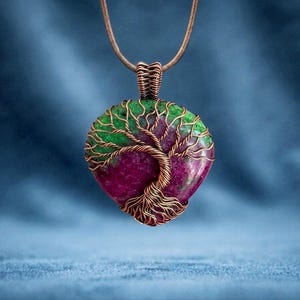 Puede incluir: Un colgante artesanal con una piedra preciosa en forma de corazón con un diseño de árbol de la vida. La piedra preciosa es una mezcla de tonos verdes y magenta, encapsulada en alambre de cobre. El colgante cuelga de un cordón de cuero marrón.