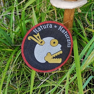 May include: A large, light brown mushroom with a white cap and a thick stem growing in a field of green grass. A black and red patch with the text "Natura + Naturans" and a white skull design is in the foreground.