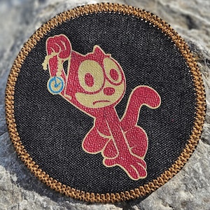 May include: A circular embroidered patch featuring a black background with a gold-colored border. The design depicts a stylized red cat character holding a small blue and gold pocket watch. The embroidery has a textured finish throughout the design.