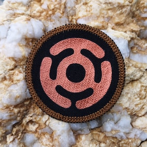 May include: A round, black and copper coloured patch with a stylised design of three overlapping circles. The patch has a brown stitched border.