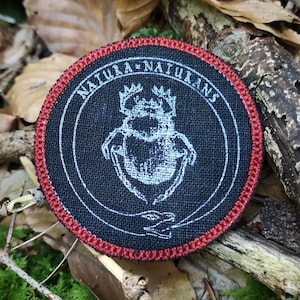 May include: A black and white embroidered patch with a red border. The patch features a beetle with a crown and the text "NATURA - NATURANS".